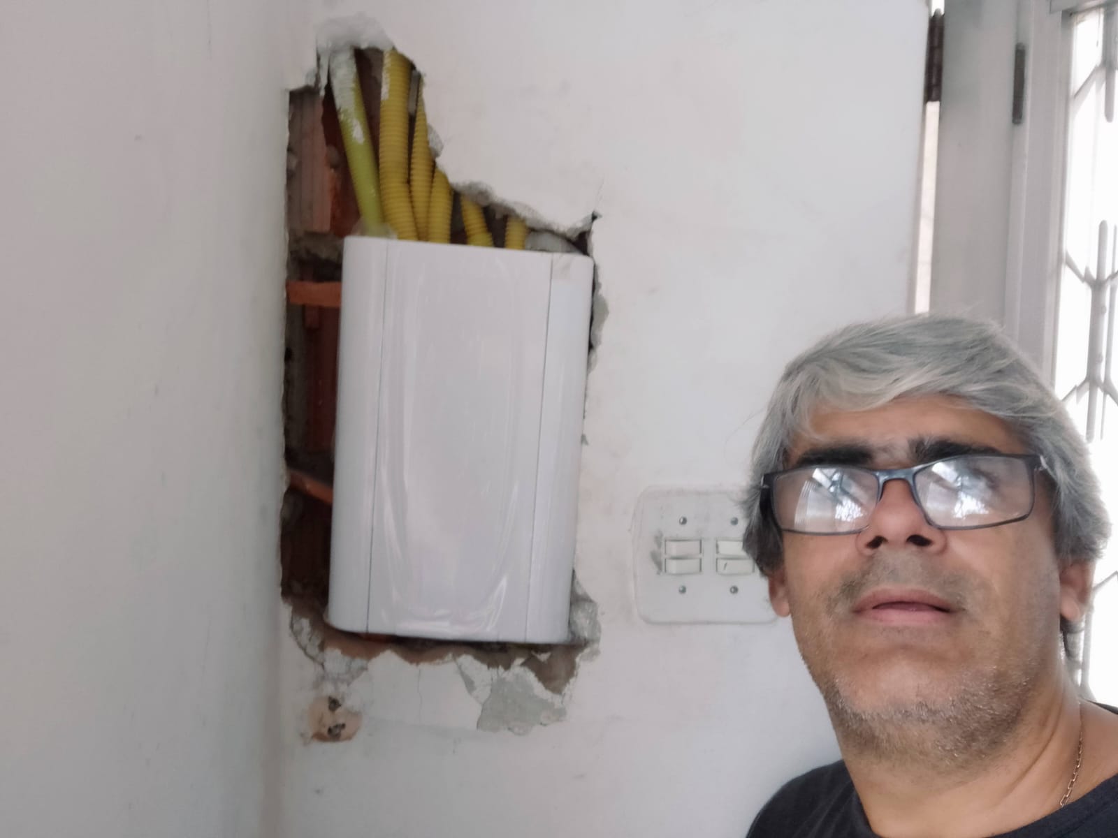 homem de óculos e cabelos grisalhos ao lado de caixa elétrica branca embutida na parede com eletrodutos amarel
